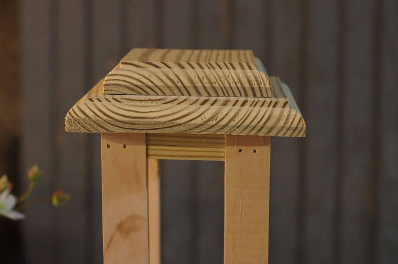 May include: A wooden lantern with a natural wood finish. The lantern has a square base and a rectangular top with a decorative, layered design.