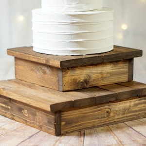 May include: Two-tiered wooden cake stand with a rustic brown finish. The stand is made of natural wood and has a simple, elegant design. It is perfect for displaying cakes, cupcakes, or other desserts.
