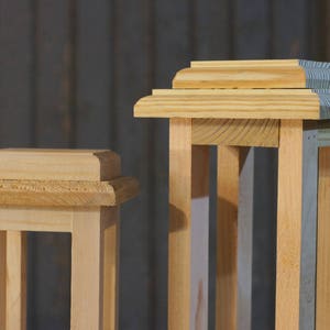 May include: Two wooden lantern frames, one with a natural wood finish and the other with a gray painted finish. Both frames have a simple, rectangular design with a decorative top.