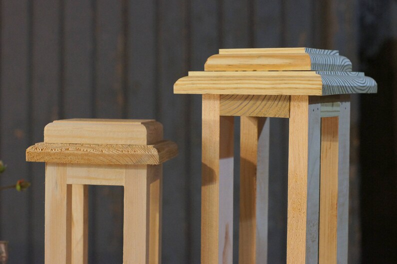 May include: Two wooden lantern frames, one with a natural wood finish and the other with a gray painted finish. Both frames have a simple, rectangular design with a decorative top.