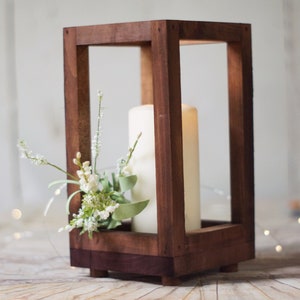 May include: A wooden candle lantern with a white pillar candle inside and a sprig of white flowers. The lantern has a dark brown stain and is made of wood.