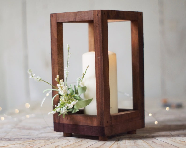 May include: A wooden candle lantern with a white pillar candle inside and a sprig of white flowers. The lantern has a dark brown stain and is made of wood.