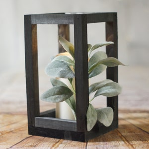 May include: A black wooden lantern with a square frame and a clear glass interior. The lantern is filled with a faux plant with green leaves.