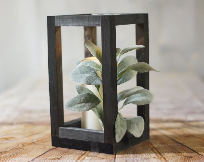 May include: A black wooden lantern with a square frame and a clear glass interior. The lantern is filled with a faux plant with green leaves.