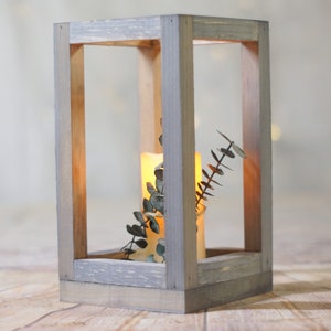 May include: A rustic wooden lantern with a gray wash finish. The lantern has a square shape and is open on all sides. A lit candle and a sprig of greenery are inside the lantern.