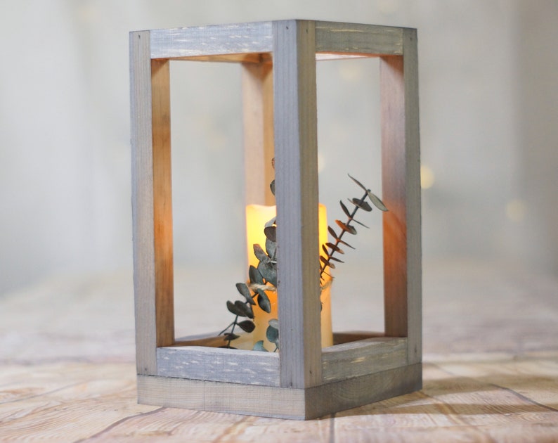 May include: A rustic wooden lantern with a gray wash finish. The lantern has a square shape and is open on all sides. A lit candle and a sprig of greenery are inside the lantern.
