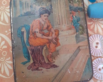 May include: Vintage tin box with a colorful illustration of a woman sitting on a chair, wearing a long orange dress and a blue sash. The box has a worn, distressed look and is likely used for storing sewing supplies.
