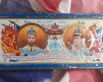 May include: A blue and white tin box with a picture of King George V and Queen Mary. The tin box is a souvenir from their visit to the Borough of Wandsworth on July 28th, 1923. The text on the tin box reads "Souvenir Their Majesties Visit to the Borough of Wandsworth" and "Copyright SNT.STA HALL".