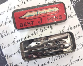 May include: A rectangular metal tin with a red lid featuring the text J CHALLENGE and BEST J PENS, containing several silver-colored metal dip pen nibs. The lid is detached and placed above the open base, which holds the metallic writing nibs.