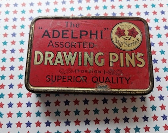 May include: A rectangular red metal tin for The Adelphi Assorted Drawing Pins. The lid features gold text reading The Adelphi Assorted Drawing Pins, Superior Quality, and a circular logo with two red ivy leaves labeled The Ivy Series.