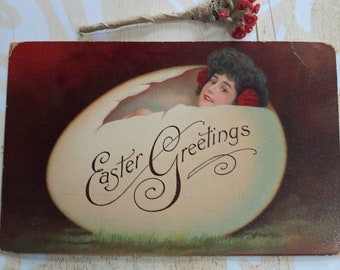 May include: Vintage Easter postcard featuring a woman with dark hair and red flowers in her hair emerging from a cracked egg. The text "Easter Greetings" is written in a decorative script.