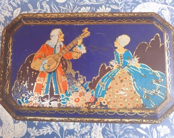 May include: Vintage rectangular metal tray with a dark blue background and a gold trim. The tray features a colorful illustration of two figures in period clothing, one playing a lute. The tray has a decorative border.