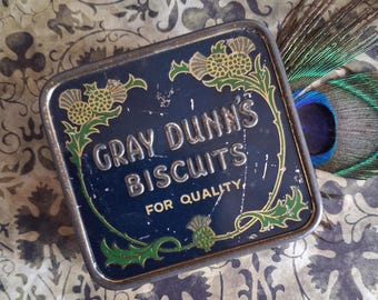 May include: A square, dark blue metal tin with rounded corners featuring the embossed text GRAY DUNN'S BISCUITS FOR QUALITY. The lid is decorated with green and gold thistle illustrations and shows signs of wear with visible surface scratches.