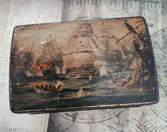 May include: A rectangular metal tin featuring a weathered illustration of the Battle of Trafalgar with sailing ships on the ocean. The top surface displays the text VICTORY AT TRAFALGAR in the upper left corner, showing signs of wear and paint loss.