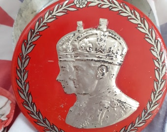 May include: A red round tin with a silver profile of a person wearing a crown. The tin is decorated with a silver laurel wreath.