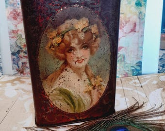 May include: Vintage metal tea tin with a red and gold design. The tin features a portrait of a woman with a floral crown and a bouquet of flowers. The tin is slightly rusted and shows signs of age.