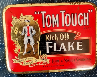 May include: A vintage tin tobacco container with a red background and gold trim. The text "Tom Tough Rich Old Flake" is printed in white and black lettering. The container features a cartoon illustration of a man smoking a pipe and the text "I smoke my pipe and sing old songs".
