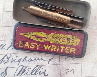 May include: An open, rectangular metal tin with a hinged lid, containing two pen nibs. The gold-colored nib is engraved with "THE LATE HINKS WELLS & CO." The tin's base is red with a yellow pen illustration and the words "MYERS EASY WRITER."