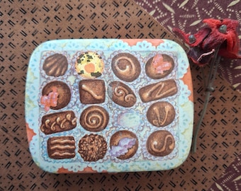 May include: A rectangular metal tin featuring a printed illustration of an assortment of chocolates in paper liners. The design includes various brown chocolate shapes with decorative swirls, some topped with pink or yellow accents.