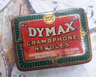 May include: A rectangular vintage tin of "DYMAX GRAMOPHONE NEEDLES" with a red background and gold lettering. The tin has a green border with a striped design and the number "200" in a circle.