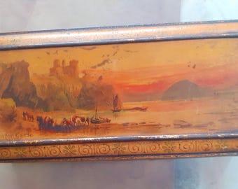 May include: Rectangular vintage tin with a landscape painting of Tintagel Castle at sunset. The painting features a castle on a cliff, a sailboat, and figures on the shore. The tin has a decorative border and the words "Tintagel Castle".