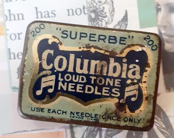 May include: A rectangular, vintage Columbia Loud Tone Needles tin. The tin is a faded green with gold and navy blue lettering. The text includes "SUPERBE", "Columbia", and "LOUD TONE NEEDLES". The tin also has the text "USE EACH NEEDLE ONCE ONLY".