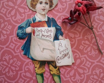 May include: A vintage-style paper cutout of a child wearing a blue jacket, yellow breeches, and green stockings, holding two white signs. One sign reads "Plenty of Cards and Presents" and the other reads "A Happy Christmas" in black script lettering.