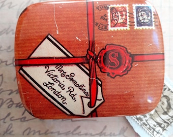 May include: A rectangular, vintage-style tin with a brown background and red ribbon design. The tin features a white tag with the text "Mrs. Semolinas, Victoria Rd, London." and a red wax seal with the letter "S". Two postage stamps are also visible.