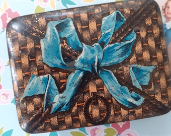 May include: A vintage tin box with a blue ribbon tied in a bow on top of a brown and orange woven pattern. The box has a rope design around the edges.