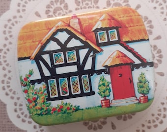 May include: A rectangular metal tin featuring a colorful illustration of a white cottage with black timber framing, a thatched roof, and a bright red door. The scene includes green bushes in pots, a small garden with red flowers, and leaded glass windows.