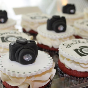 May include: A close-up of red velvet cupcakes decorated with white frosting and black fondant cameras. Some cupcakes have the word "Smile" written on them.