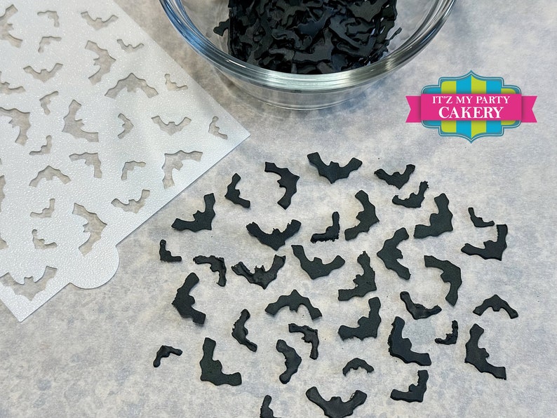 May include: A white stencil with a repeating pattern of bats, next to a bowl of black fondant bats. The bowl is on a white surface. The text "IT'Z MY PARTY CAKERY" is in the upper right corner of the image.