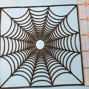 May include: Black spiderweb stencil with a square shape and a detailed design. The stencil is approximately 5 inches wide.