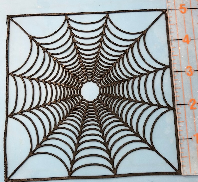 May include: Black spiderweb stencil with a square shape and a detailed design. The stencil is approximately 5 inches wide.
