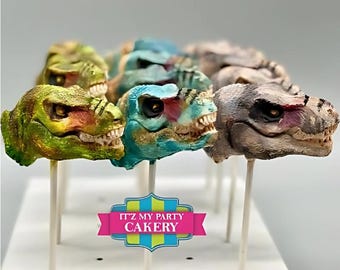 May include: A collection of colorful dinosaur head cake pops on sticks. The cake pops are decorated with realistic details and are arranged on a white surface. The text "ITZ MY PARTY CAKERY" is visible in the lower center of the image.