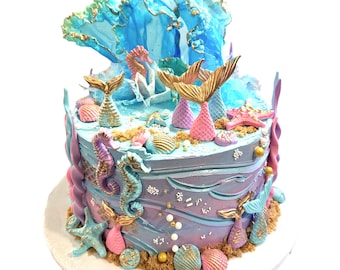 May include: A blue and purple mermaid-themed cake decorated with seashells, starfish, seahorses, and mermaid tails. The cake is topped with a blue and green wave.