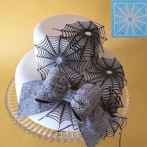 May include: A two-tiered white cake decorated with black icing in a spiderweb design. A gray lace bow is placed on the bottom tier of the cake.
