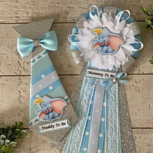 May include: Blue and silver baby shower decorations with a Dumbo theme. The decorations include a tie with a bow tie and a pin with a ribbon. The tie says "Daddy To Be" and the pin says "Mommy To Be".