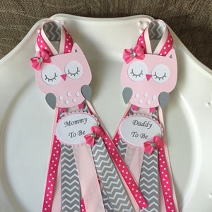May include: Two pink and gray owl-shaped baby shower pins with pink and gray ribbons. The pins say "Mommy to Be" and "Daddy to Be".