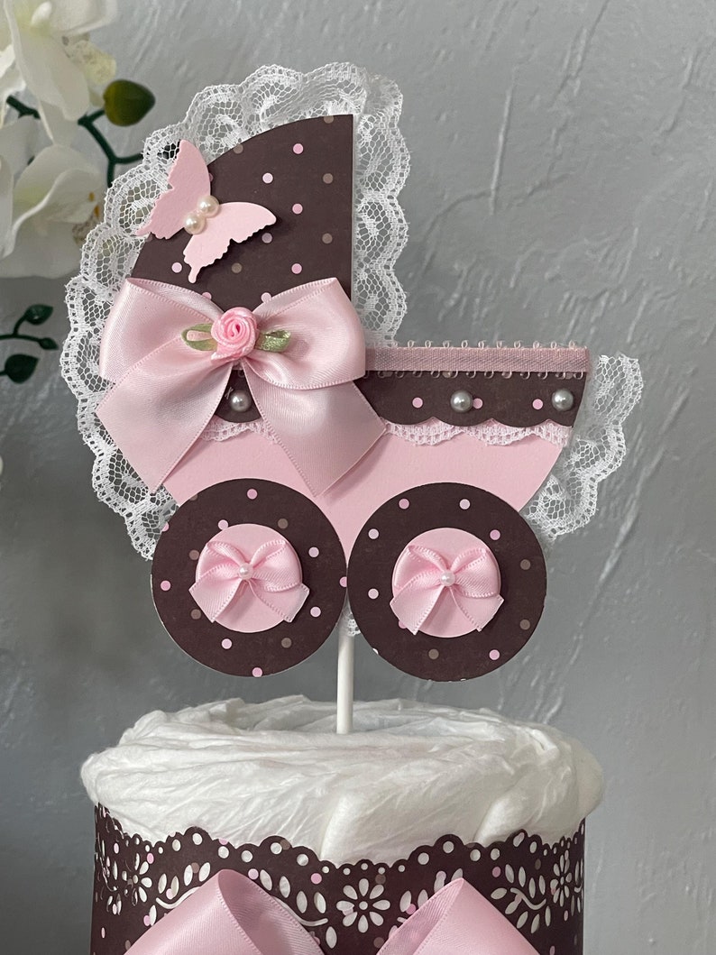 Peut inclure: Un g&acirc;teau de couches en forme de landau b&eacute;b&eacute;, de couleur marron et rose, avec un n&oelig;ud rose, un papillon rose et de la dentelle blanche. Le g&acirc;teau est d&eacute;cor&eacute; de pois marrons et de perles blanches.
