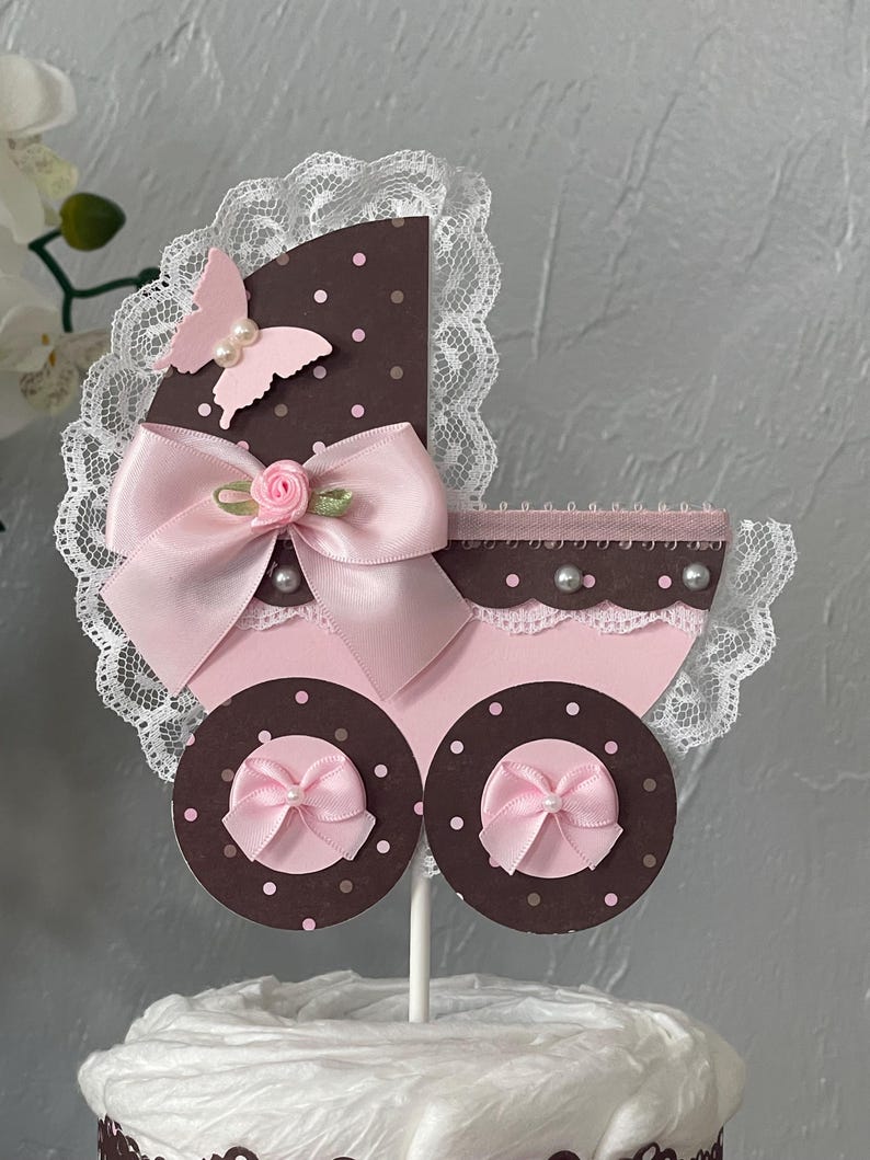 Peut inclure: Un topper de g&acirc;teau de chariot de b&eacute;b&eacute; rose et marron avec un n&oelig;ud rose, un papillon rose et une bordure en dentelle blanche. Le chariot est d&eacute;cor&eacute; de pois blancs.