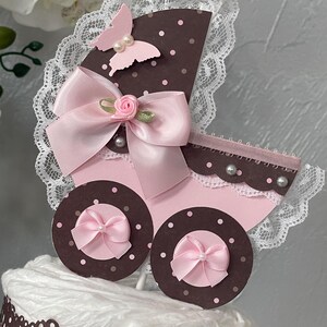 Peut inclure: Un g&acirc;teau de couches pour b&eacute;b&eacute; en forme de landau rose et marron avec un n&oelig;ud rose, un papillon rose et une bordure en dentelle blanche.