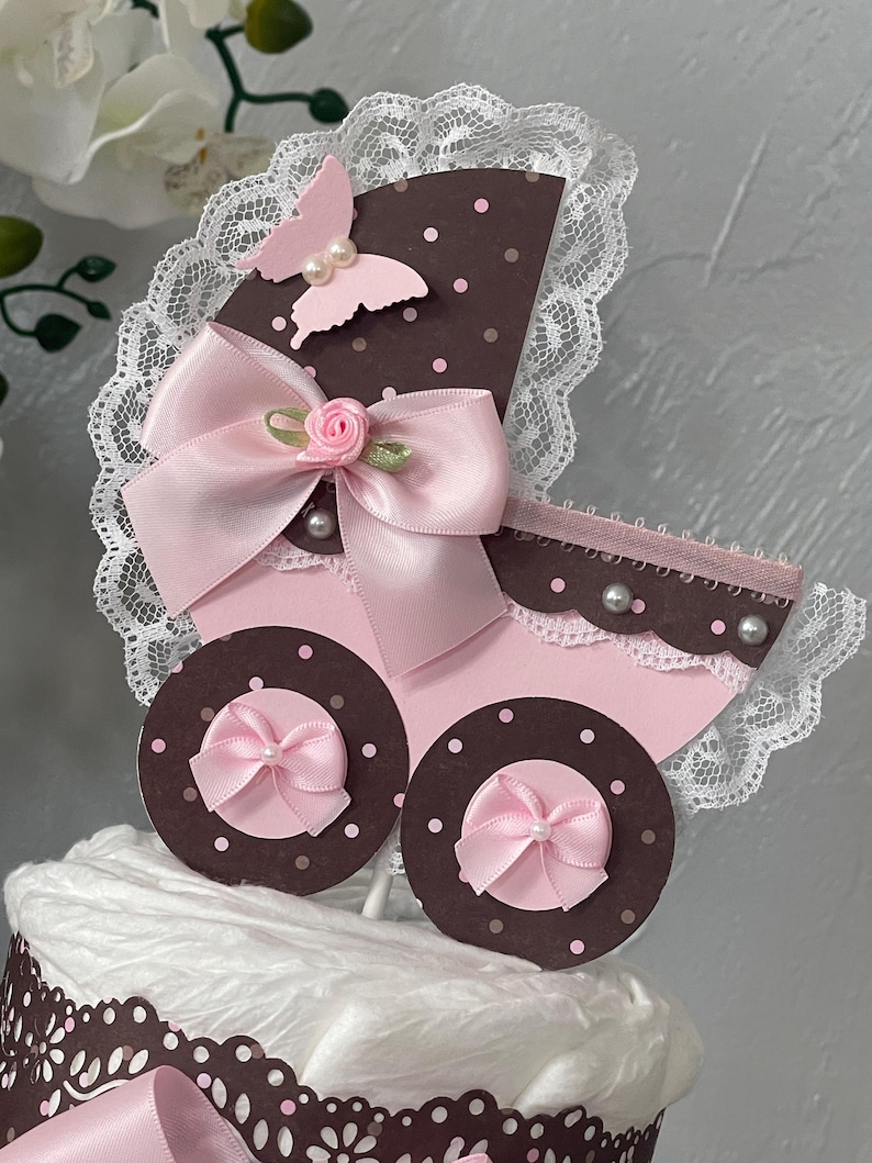 Peut inclure: Un g&acirc;teau de couches pour b&eacute;b&eacute; en forme de landau rose et marron avec un n&oelig;ud rose, un papillon rose et une bordure en dentelle blanche.