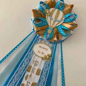May include: Blue and gold baby shower sash with a hot air balloon design and the text "Mommy To Be" and "Hello Little One".