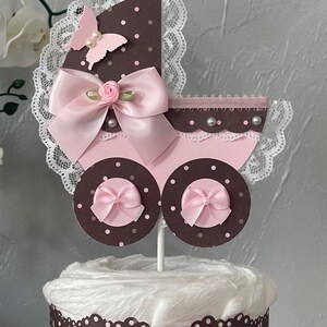 Peut inclure: Un g&acirc;teau de couches en forme de poussette de b&eacute;b&eacute; rose et marron avec un n&oelig;ud rose, de la dentelle blanche et un papillon rose. Le g&acirc;teau est d&eacute;cor&eacute; de pois blancs et de perles.