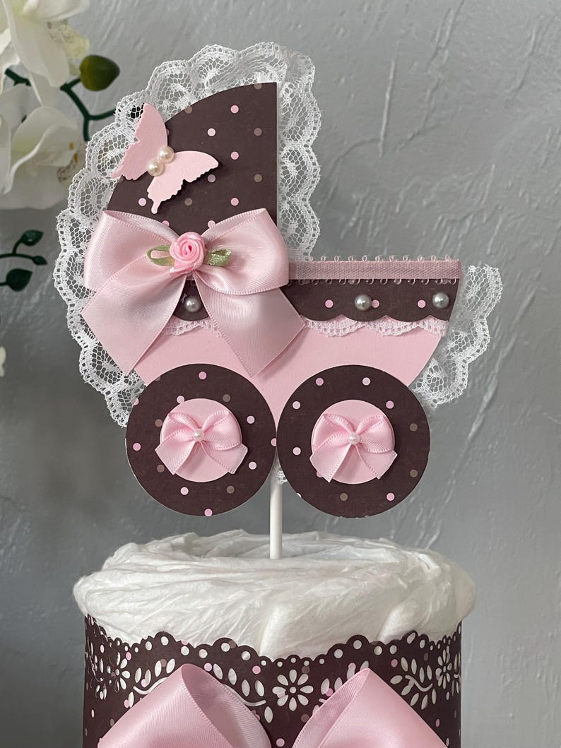 Peut inclure: Un g&acirc;teau de couches en forme de poussette de b&eacute;b&eacute; rose et marron avec un n&oelig;ud rose, de la dentelle blanche et un papillon rose. Le g&acirc;teau est d&eacute;cor&eacute; de pois blancs et de perles.