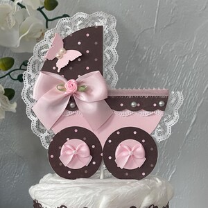 Peut inclure: Un g&acirc;teau de couches marron et rose avec un dessus de poussette rose et blanc. Le dessus de poussette a un n&oelig;ud rose, un papillon rose et des pois blancs.