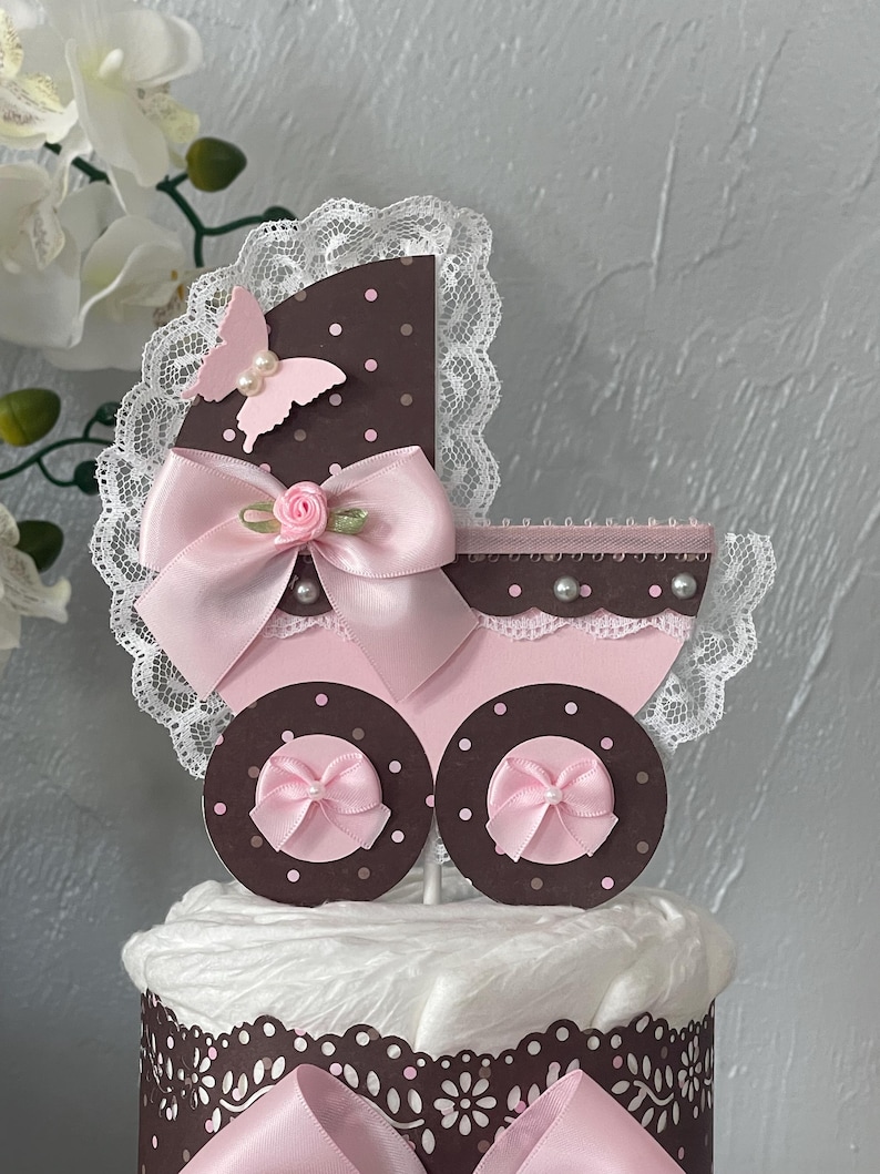 Peut inclure: Un g&acirc;teau de couches marron et rose avec un dessus de poussette rose et blanc. Le dessus de poussette a un n&oelig;ud rose, un papillon rose et des pois blancs.