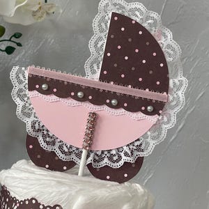 Peut inclure: Un topper de g&acirc;teau de chariot de b&eacute;b&eacute; rose et marron avec une bordure en dentelle blanche et des strass. Le chariot est sur un b&acirc;ton et est pr&ecirc;t &agrave; &ecirc;tre plac&eacute; sur un g&acirc;teau.