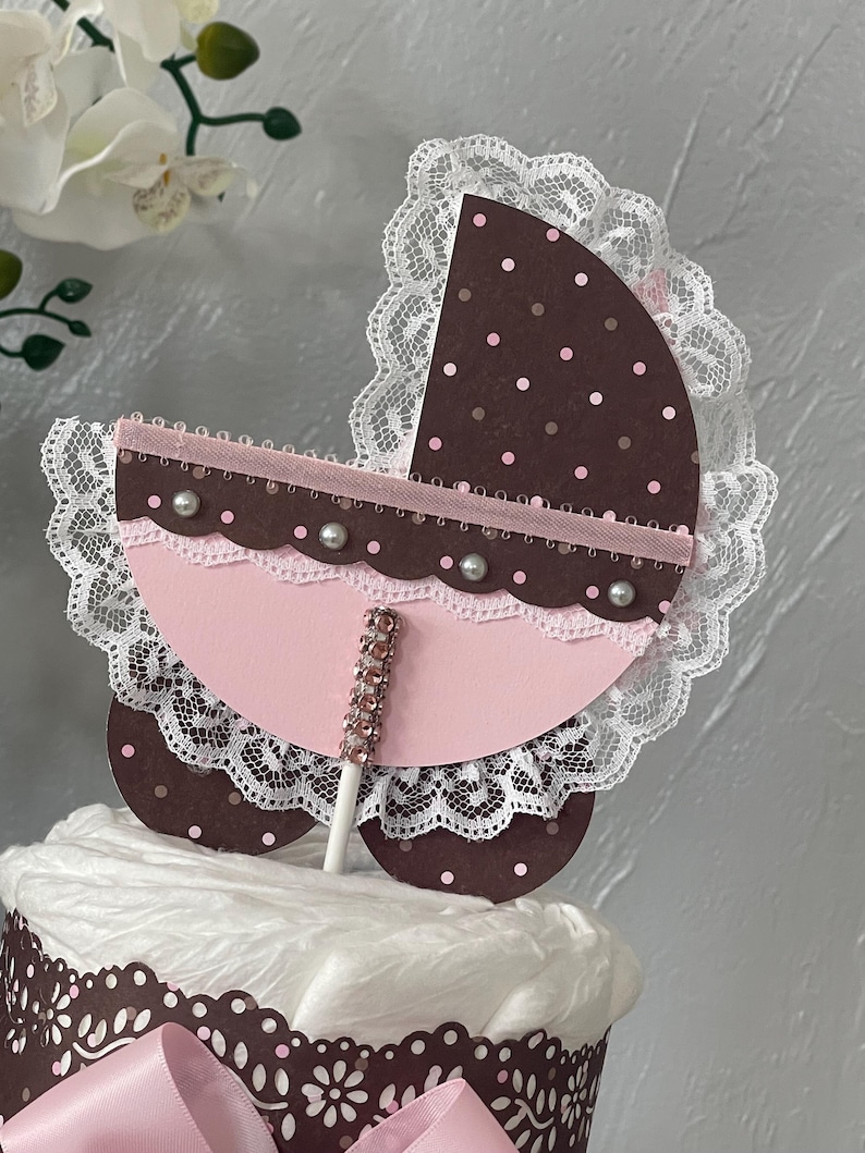 Peut inclure: Un topper de g&acirc;teau de chariot de b&eacute;b&eacute; rose et marron avec une bordure en dentelle blanche et des strass. Le chariot est sur un b&acirc;ton et est pr&ecirc;t &agrave; &ecirc;tre plac&eacute; sur un g&acirc;teau.